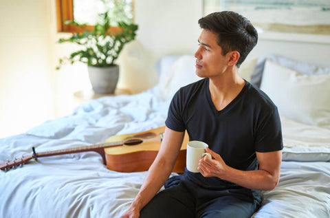 A young man sitting on the edge of the bed and holding a cup of coffee while wearing a Slumber Cloud UltraCool V-Neck Tee