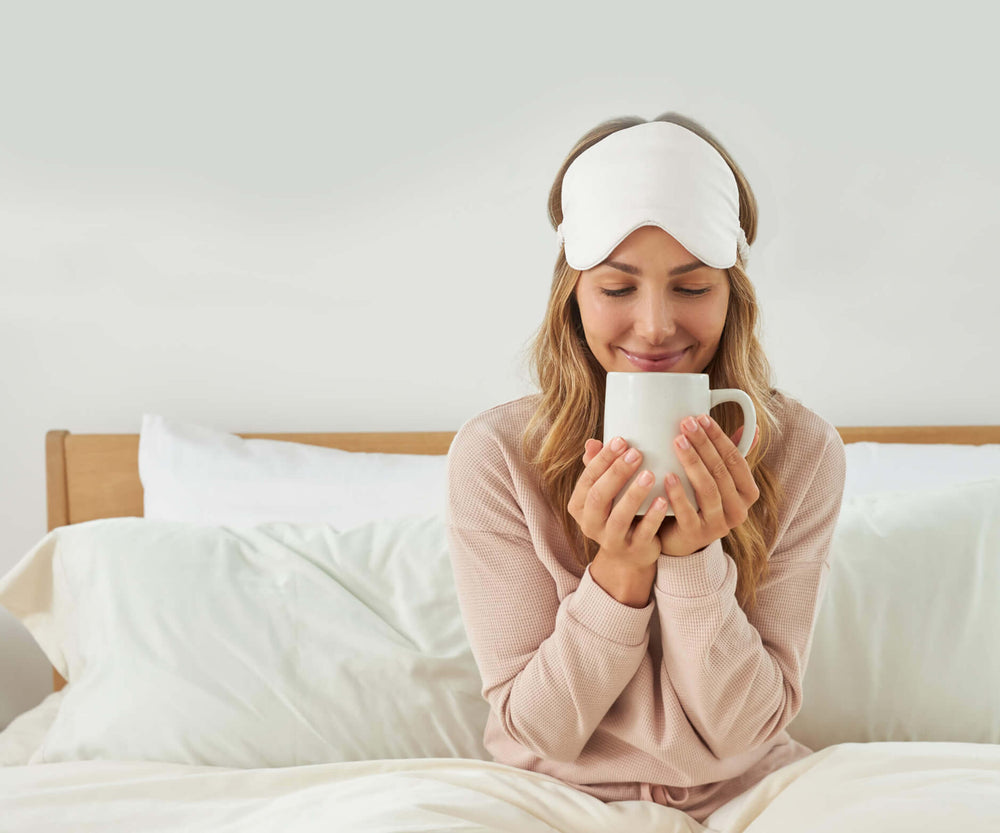 A woman smiling and drinking a cup of coffee while sitting in a bed made with Slumber Cloud products