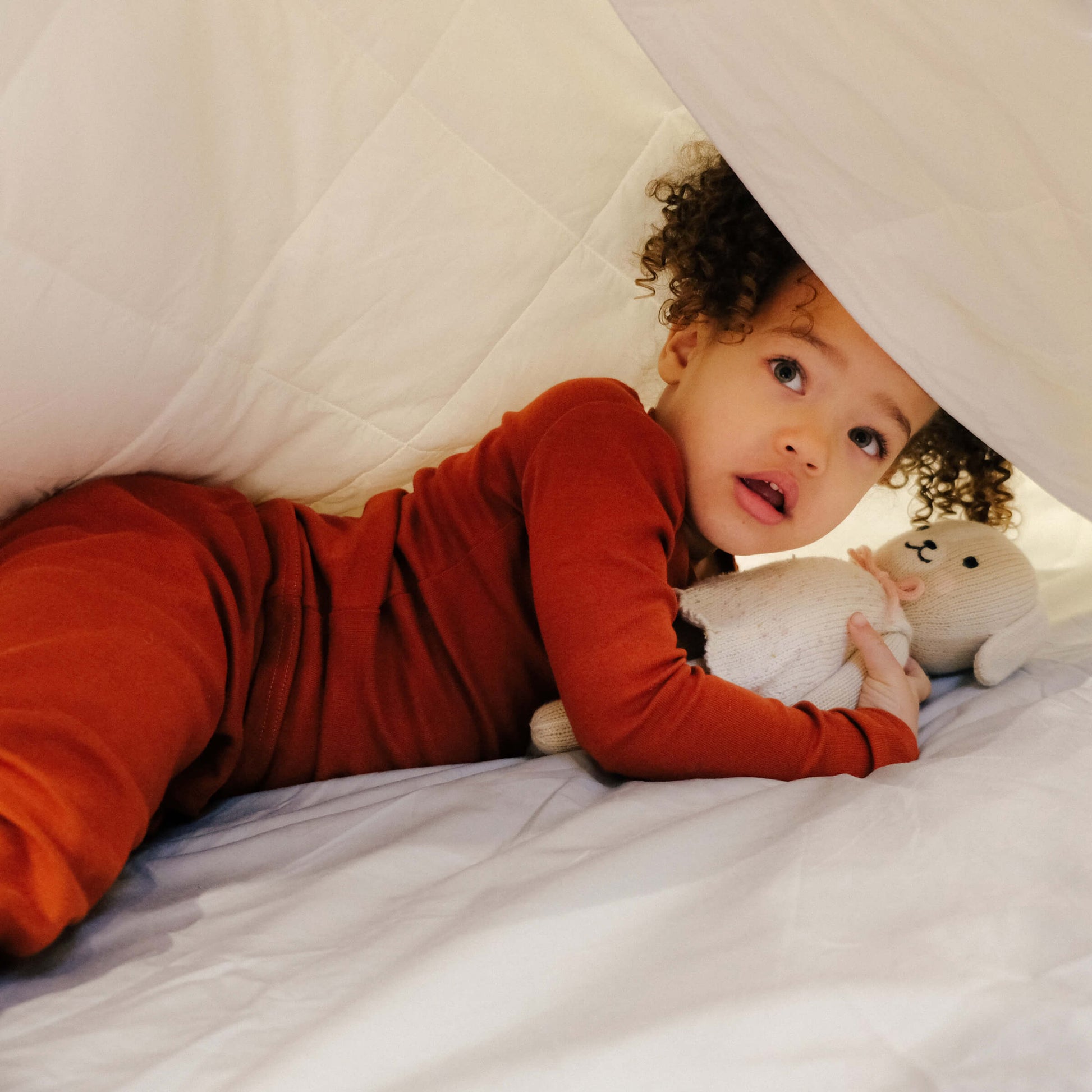 A child holding a teddy bear lying under the Slumber Cloud Comforter made with temperature regulation technology